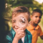 Barn i naturen där en av pojkarna tittar glatt igenom ett förstoringsglas